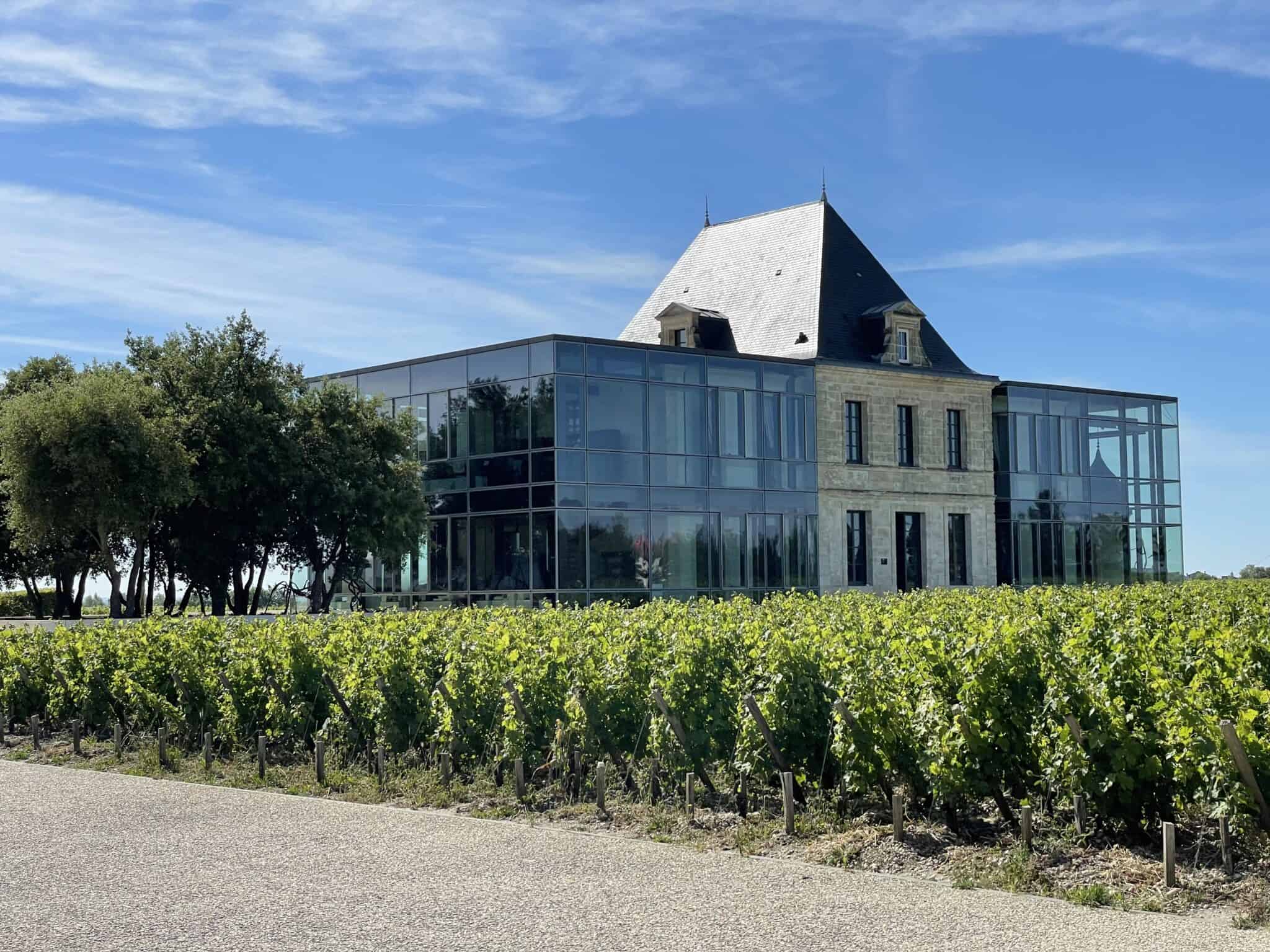Château Pédesclaux à Pauillac, Grand Cru Classé du Médoc, bâtiment moderne avec façades en verre et section centrale en pierre, entouré de vignobles verdoyants sous un ciel bleu, dans la région viticole de Bordeaux.