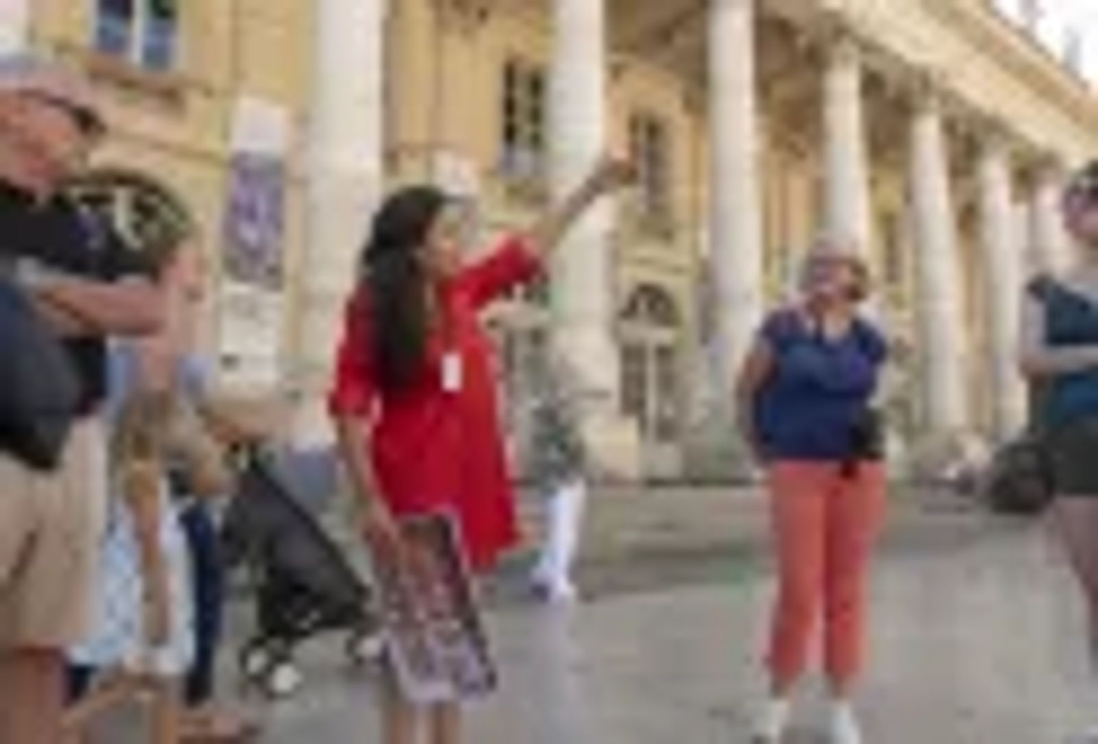 Une guide touristique en robe rouge fait une visite guidée devant le Grand Théâtre de Bordeaux, en montrant l'architecture classique du bâtiment à un groupe de visiteurs comprenant des enfants et des adultes.